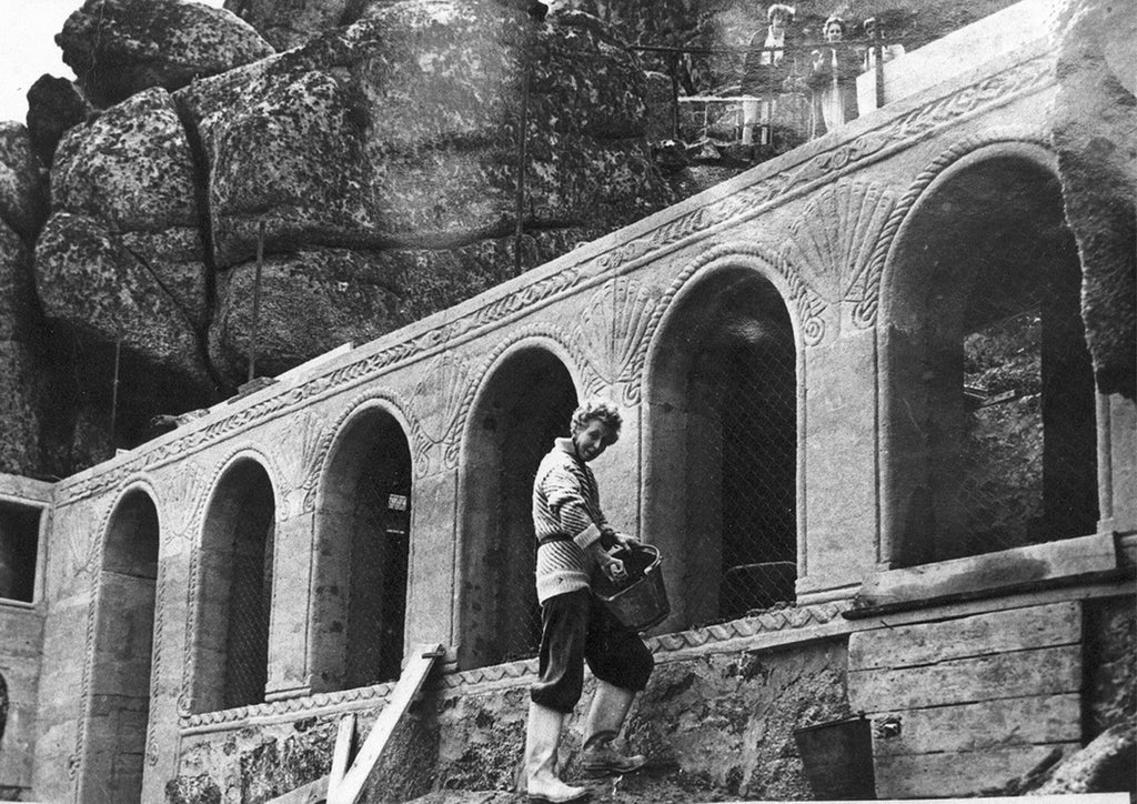 This is a black and white image and shows Cade working on the stage for the Minack Theatre. In the foreground, she is wearing dark waterproof trousers and light knee-high wellington boots. She is wearing a long black and white striped woollen cardigan and is slightly rolled up at the elbows. Cade is perched on some rugged and unfinished ground holding a black bucket and is seemingly mixing cement by hand. In the background are concrete pillars engraved with scallops, with waves along the trims. 