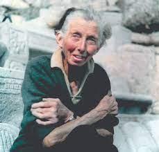 This photograph shows Rowena Cade in the later years of her life. She is wearing a dark green V-neck jumper with a white collared shirt layered beneath; her hair is grey, short and wavy, it is tied up and appears unruly in the coastal wind. Cade is hugging her arms to her chest and smiling towards the left, as she faces the sunshine. In the background, are the granite boulders of the Minack Theatre.