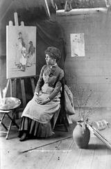 A photograph of Elizabeth Forbes in her studio, sitting next to an easel with an unfinished painting on it.