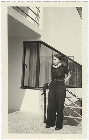 A photograph of Bryher at Kenwin, their house in Switzerland. They have short, dark hair and are wearing a short sleeved, belted jumper over a white shirt and wide-legged trousers. They are leaning against a railing. and looking to the left of the picture.